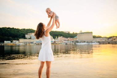 çocuğu gün batımına ve suya karşı döndüren anne. Mutlu anne ve bebek. Sahilde oynuyorum. Oğlunu havaya kaldıran genç kadın