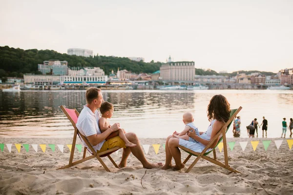 Family at seaside in evening open-air cafe. Mother and father and two sons sit on sun loungers, looking at sunset on sandy beach near river overlooking city. Concept travel and summer family vacation