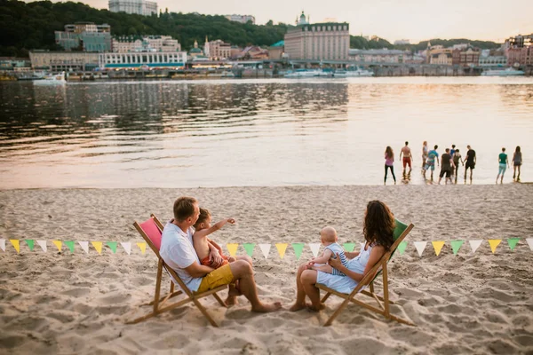Family at seaside in evening open-air cafe. Mother and father and two sons sit on sun loungers, looking at sunset on sandy beach near river overlooking city. Concept travel and summer family vacation