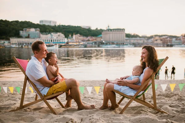 Family at seaside in evening open-air cafe. Mother and father and two sons sit on sun loungers, looking at sunset on sandy beach near river overlooking city. Concept travel and summer family vacation