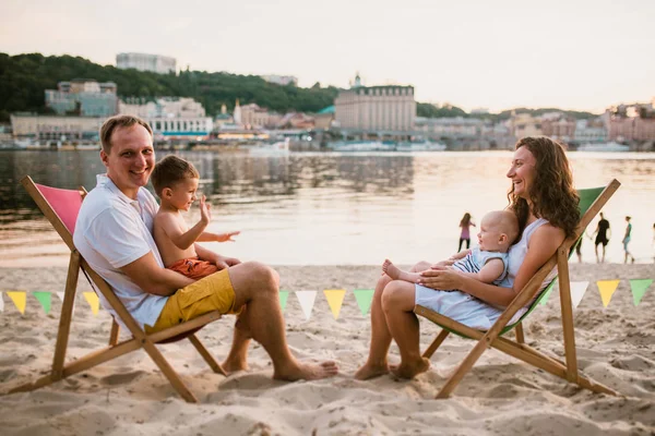 Familia en la playa en la cafetería al aire libre por la noche. Madre y padre y dos hijos se sientan en tumbonas, mirando la puesta de sol en la playa de arena cerca del río con vistas a la ciudad. Concepto de viaje y vacaciones familiares de verano —  Fotos de Stock