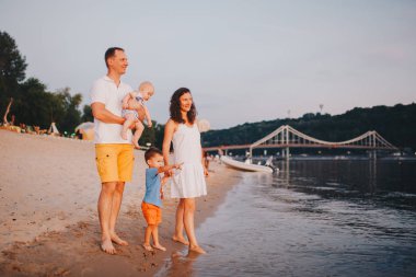 Yaz aylarında Nehir tarafından Açık Havada İki Toddler Çocuk lu Aile Görünümü. Mutlu genç aile gün batımında sahilde eğlenin. Aktif ebeveynler ve çocuklarla yaz tatillerinde insanlar açık hava etkinliği