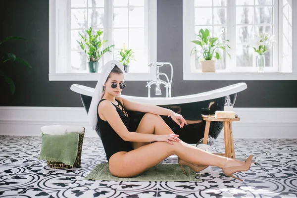 Theme fashion and style. Holiday rich women in spa expensive hotel. Caucasian young sexy girl sits on floor near bathroom in black swimsuits and high heel shoes. bathroom interior — Stock Photo, Image