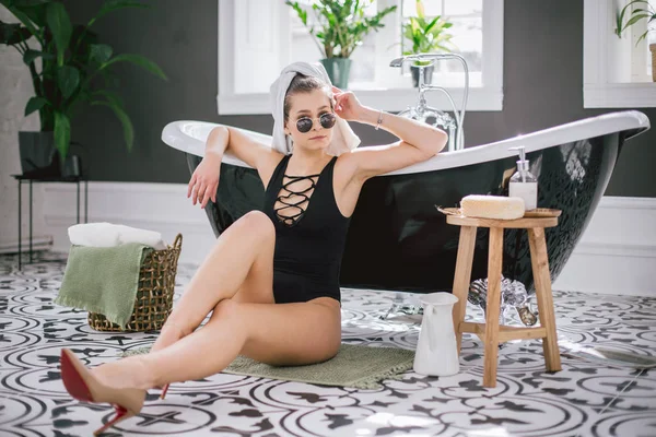 Thema Mode und Stil. Urlaub reichen Frauen in Spa-Hotel teuer. Kaukasische junge sexy Mädchen sitzt auf dem Boden neben Badezimmer in schwarzen Badeanzügen und Stöckelschuhen. Badezimmerausstattung — Stockfoto
