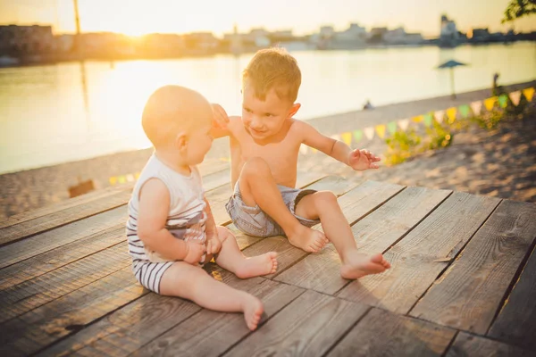 川岸の桟橋に座っている二人の小さな男の子。友情と友愛の概念。晴れた楽しい夏の日。桟橋に座っている子供たち。木製の桟橋に座っている異なる年齢の2人の子供 — ストック写真