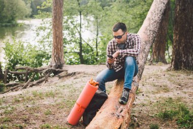 Genç adam ormanda kamyonda oturup cep telefonu kullanıyor. adam gezgin elinde telefon tutarak, büyük düşmüş ağaç üzerinde oturur. tema yürüyüş doğa seyahat. Açık hava etkinlikleri yürüyüş. Seyahat ve macera