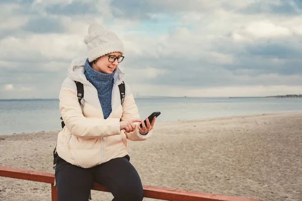 Donna caucasica in cappello e giacca con uno zaino in inverno si siede su un molo di legno sulla spiaggia vicino al Mare del Nord. Danimarca Copenhagen turista utilizza un telefono cellulare sulla riva. Tema del viaggio in Scandinavia — Foto Stock