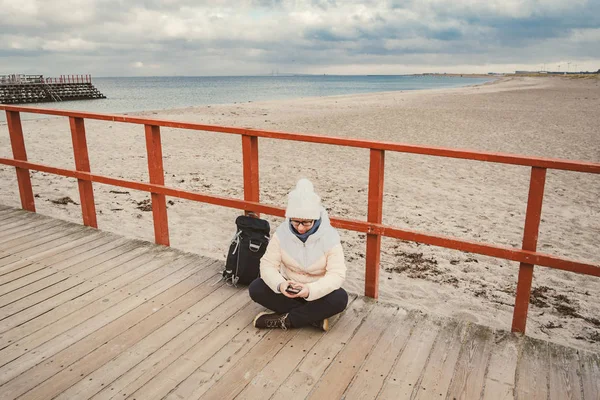 Белая женщина в шляпе и пиджаке с рюкзаком зимой сидит на деревянном пирсе на пляже у Северного моря. Дания Копенгаген турист использует мобильный телефон на берегу. Скандинавия Трэвел — стоковое фото