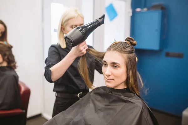 Portret van gelukkige vrouw in de kapsalon. Professioneel haar styling concept. Kapper drogen meisje lang haar met behulp van haardroger en borstel. Drogen met föhn — Stockfoto