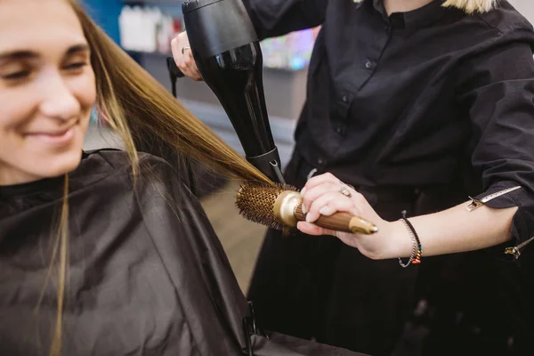 Портрет счастливой женщины в парикмахерской. Концепция укладки волос. Парикмахер сушит девушке длинные волосы с помощью фена и щетки. Сушка с сушилкой — стоковое фото