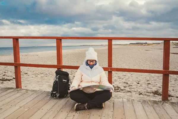 Кавказька жінка в капелюсі і кофта з рюкзаком взимку сидить на дерев'яному пірсі на пляжі біля Північного моря. Данія Копенгаген турист використовує папери карта берега. Тематичні подорожі та навігація в Європі — стокове фото
