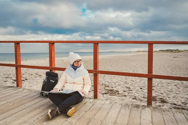 Donna caucasica in cappello e giacca con zaino in inverno si siede sul molo di legno sulla spiaggia vicino al Mare del Nord. Danimarca Il turismo di Copenaghen utilizza mappe cartacee. Tema Viaggi e navigazione in Europa — Foto Stock