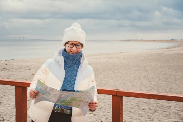 Femme caucasienne en chapeau et veste avec sac à dos en hiver se trouve sur une jetée en bois sur la plage près de la mer du Nord. Danemark Copenhague touriste utilise papier carte rivage. Thème Voyage et navigation en Europe — Photo