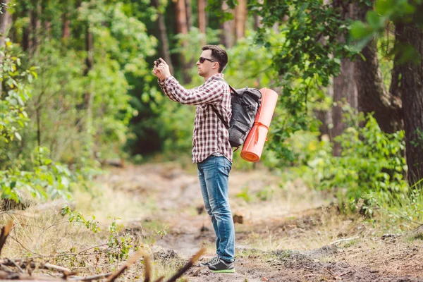 Man vandrare tar foto smart telefon i skogen. Attraktiv resenären gör foto med mobiltelefon. Traveler vandrare man med ryggsäck vandring i naturen, turist Backpacker använda kameran. Vacation Concept — Stockfoto