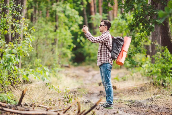 Man vandrare tar foto smart telefon i skogen. Attraktiv resenären gör foto med mobiltelefon. Traveler vandrare man med ryggsäck vandring i naturen, turist Backpacker använda kameran. Vacation Concept — Stockfoto