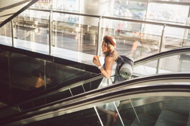 Yürüyen Merdiven'de Havaalanında Şapka Takan Portre Gülümseyen Kadın. el bagajı ile seyahat eden insanlar. Tema turizm ve ulaşım. Bir yürüyen merdiven üzerinde havaalanı terminalinde kahve ile Kafkas kız