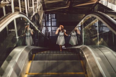 Yürüyen Merdiven'de Havaalanında Şapka Takan Portre Gülümseyen Kadın. el bagajı ile seyahat eden insanlar. Tema turizm ve ulaşım. Bir yürüyen merdiven üzerinde havaalanı terminalinde kahve ile Kafkas kız