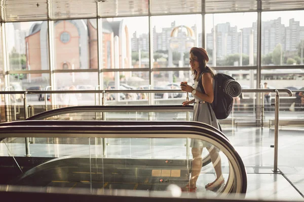 Retrato sonriente mujer con sombrero en el aeropuerto en escaleras mecánicas. personas que viajan con equipaje de mano. Turismo temático y transporte. Chica caucásica con café en la terminal del aeropuerto en una escalera mecánica — Foto de Stock