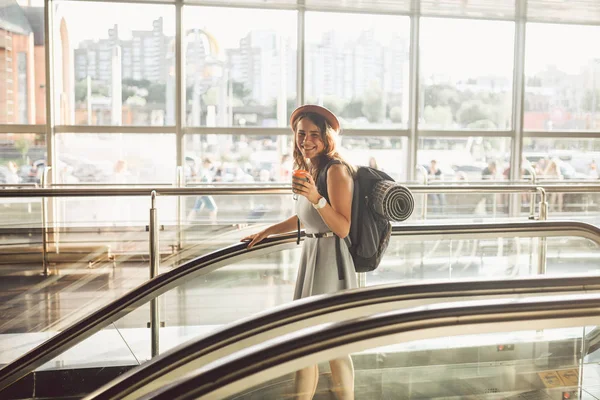 Портрет усміхнена жінка носить капелюх в аеропорту на ескалаторі. люди, які подорожують з ручним багажем. Тематичний туризм і транспорт. Кавказька дівчина з кавою в терміналі аеропорту на ескалаторі — стокове фото