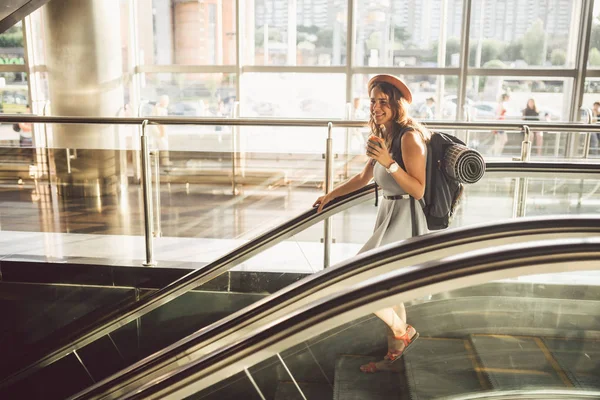 Retrato Sorrindo Mulher vestindo chapéu no aeroporto em escada rolante. pessoas que viajam com bagagem de mão. Turismo temático e transporte. Menina caucasiana com café no terminal do aeroporto em uma escada rolante — Fotografia de Stock