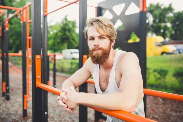 Portrait young bearded man standing on public sports ground and training on parallel bars. Handsome workout athlete. Portrait sporty man with beard, rest after training. Summer workout outdoors — Stock Photo, Image