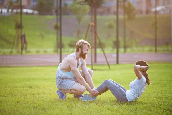 Sit UPS fitness pár gyakorlása ülni kint a fűben. Fit emberek dolgoznak ki kereszt képzést. Nő csinál hasi crunches sajtó gyakorlás-val edző. Pár doing Sit-UPS hasi összeroppant — Stock Fotó