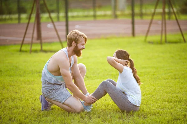 Sit ups fitness couple exercising sit up outside in grass. Fit people working out cross training. Woman doing abdominal crunches press exercise with trainer. Couple Doing Sit-Ups Abdominal Crunch