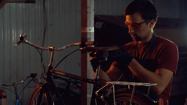 theme small business bike repair. A young Caucasian brunette man wearing safety goggles, gloves and an apron uses a hand tool to repair and adjust the bike in the workshop garage