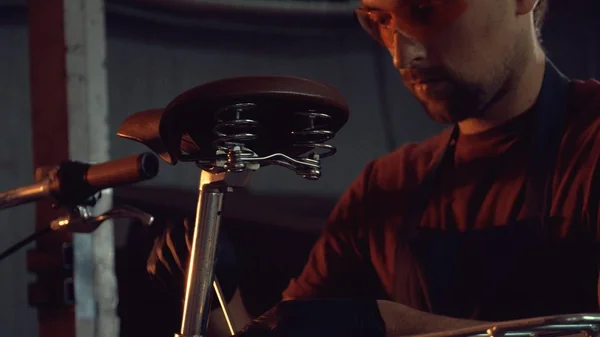 theme small business bike repair. A young Caucasian brunette man wearing safety goggles, gloves and an apron uses a hand tool to repair and adjust the bike in the workshop garage