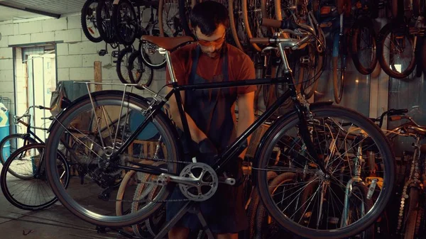 Thema Reparatur von Geschäftsfahrrädern. Junge kaukasische brünette Mann mit Schutzbrille, Handschuhen und Schürze benutzt Handwerkzeug Reparatur und Einstellung Kurbel und Pedalsystem Fahrrad in der Werkstatt — Stockfoto