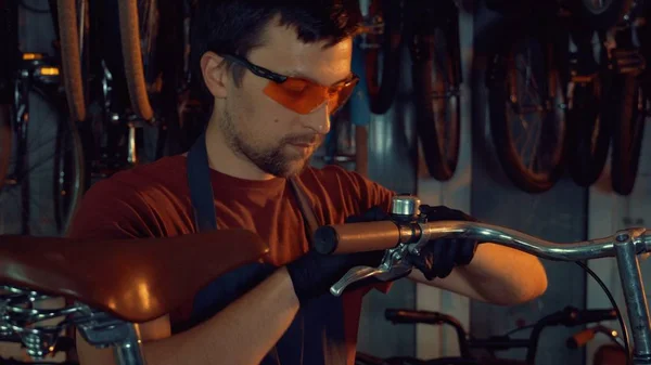 theme small business bike repair. A young Caucasian brunette man wearing safety goggles, gloves and apron uses hand tool to repair and adjust the Handbrabar brake bike handle in garage of workshop