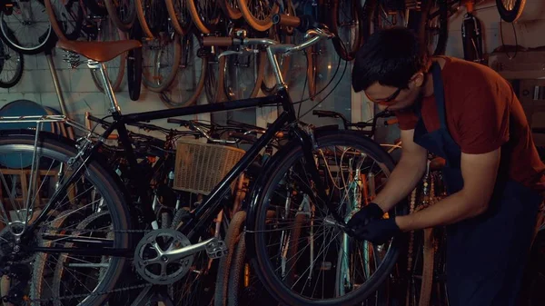 theme small business bike repair. Young brunette Caucasian man in protective glasses, gloves and fartuhe uses hand tools to repair and tuning Rim Brakes and wheel spinning bike in a garage workshop