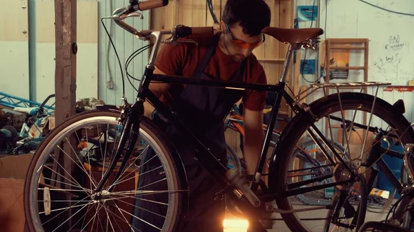 Temă repararea bicicletelor de afaceri mici. Un tânăr brunetă caucaziană purtând ochelari de siguranță, mănuși și un șorț insistă asupra întrerupătoarelor de viteză de pe ghidonul bicicletei din garajul atelierului — Fotografie, imagine de stoc