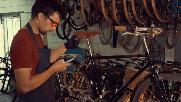 theme small business bike repair. Young caucasian brunette man wearing safety goggles, gloves and fartukhe uses mobile phone technology, takes notes, checklist in bicycle workshop