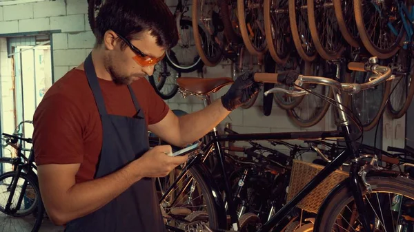 theme small business bike repair. Young caucasian brunette man wearing safety goggles, gloves and fartukhe uses mobile phone technology, takes notes, checklist in bicycle workshop