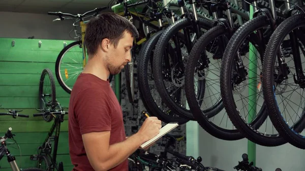 küçük iş bisiklet satış Tema. Genç beyaz erkek esmer küçük işletme sahibi, mağaza yöneticisi yapar notları, not defteri ve kalem Bisiklet mağazasında denetim listesi kullanır