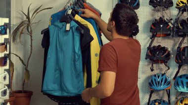 Genç bir beyaz adam yakınındaki bir raflar giysiler bir askı bir bisiklet mağazası üzerinde Bisiklete binme spor duruyor. Bir bisiklet giysiler seçmek ve kask