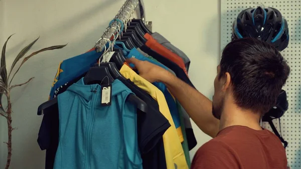 A young Caucasian man is standing near a racks of sports cycling clothes on a hanger in a bicycle store. Choosing a bicycle clothes and helmet