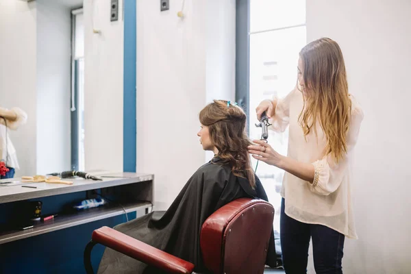 Beleza, conceito de penteado, mulher jovem feliz e cabeleireiro com ferro de cabelo fazendo penteado no salão de cabeleireiro. Mulher com cabelo cabeleireiro estilizado. Suaviza o encrespamento do cabelo. Stylist Usando Ferramenta para Modelagem — Fotografia de Stock