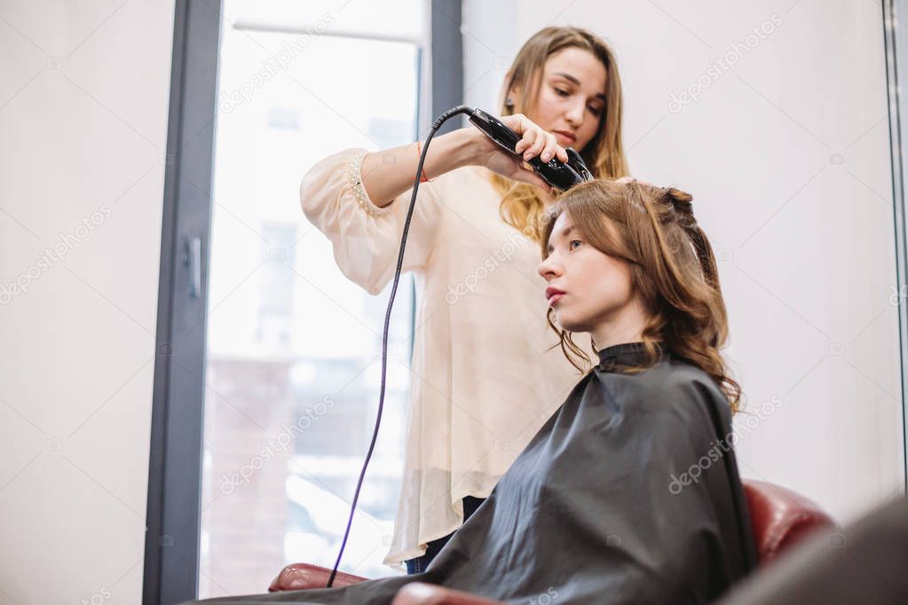 beauty, hairstyle concept, happy young woman and hairdresser with hair iron making hairdo at hair salon. Woman Having Hair Stylized Hairdresser. Smoothes Hair Curling. Stylist Using Tool for Modeling