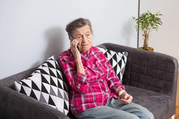 An old elderly woman grandmother with gray hair sits at home on the couch using the hand phone, a telephone conversation to hear the bad news. Emotion fear scare