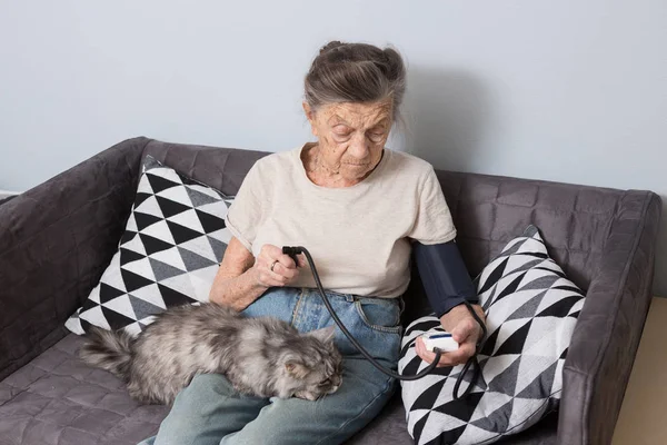 The topic is very old person and health problems. A senior Caucasian woman, 90 years old, with wrinkles and gray hair, sits home on sofa with pet cat and uses tonometer. High blood pressure headache
