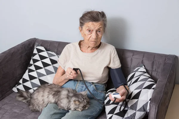 The topic is very old person and health problems. A senior Caucasian woman, 90 years old, with wrinkles and gray hair, sits home on sofa with pet cat and uses tonometer. High blood pressure headache