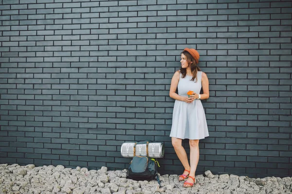 Chica alegre en vestido sosteniendo llevar taza. Hipster modelo con elegante traje de verano al aire libre. mujer con mochila pasea por la calle de la ciudad. chica hipster en la pared de ladrillo, estilo de vida de la adolescencia moderna — Foto de Stock