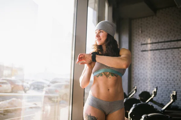 Tema do esporte e tecnologia. Uma mulher forte e bonita com uma barriga plana perto da janela em um dia ensolarado aperta os botões, usa um relógio de esportes preto em seu braço. Em linho cinza, calças curtas e um chapéu — Fotografia de Stock