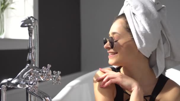 Молодая белая женщина с полотенцем на голове и солнцезащитными очками, проходящая процедуру в салоне красоты, во внутренней комнате. Отдых в ванной комнате в постельном белье. Концепция ухода за телом и расслабления — стоковое видео