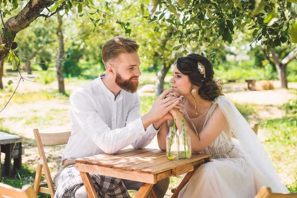 Casal atraente recém-casados, momento feliz e alegre. noiva e noivo sentam-se à mesa para dois na floresta. Conceito encontro romântico. Casamento casal sentado na mesa de café e amorosamente olhar um para o outro — Fotografia de Stock