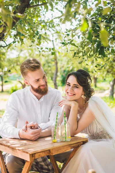 Atractiva pareja de recién casados, momento feliz y alegre. novia y el novio se sientan a la mesa para dos en el bosque. Concepto de cita romántica. Pareja de boda sentada en la mesa de la cafetería y amorosamente se miran —  Fotos de Stock