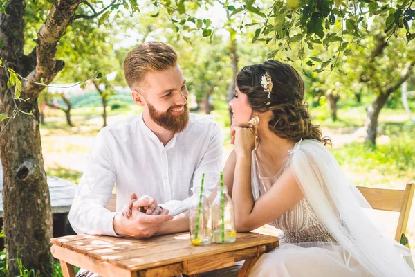 Çekici çift yeni evliler, mutlu ve neşeli an. gelin ve damat ormanda iki için belirlenen masada oturup. Kavram romantik tarih. Düğün çift kafe masada oturan ve sevgiyle birbirlerine bakmak