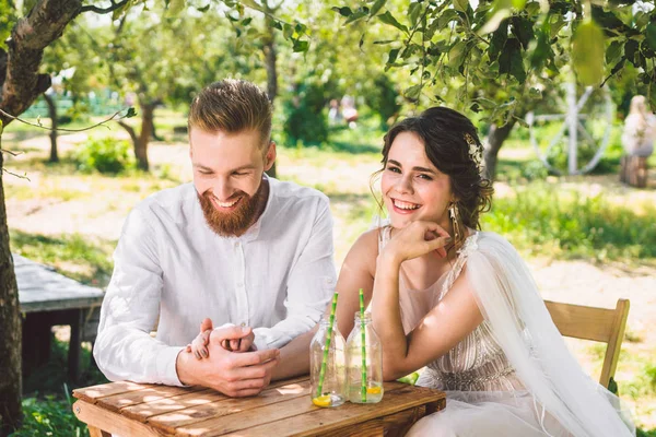 Casal atraente recém-casados, momento feliz e alegre. noiva e noivo sentam-se à mesa para dois na floresta. Conceito encontro romântico. Casamento casal sentado na mesa de café e amorosamente olhar um para o outro — Fotografia de Stock